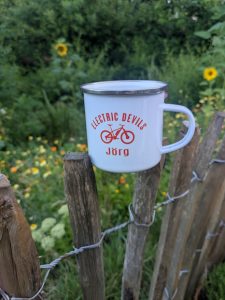 weiße Emaille Tasse der Electric Devils mit Namen "Jörg" darauf, stehend auf dem Gartenzaun voe der Blühwiese in unserem Garten. 
